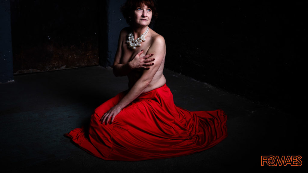 Elegante, reife Dame in stilvollem Ambiente. Mit Perlenkette und rotem Rock in dunkler Umgebung. Sitzend am Boden