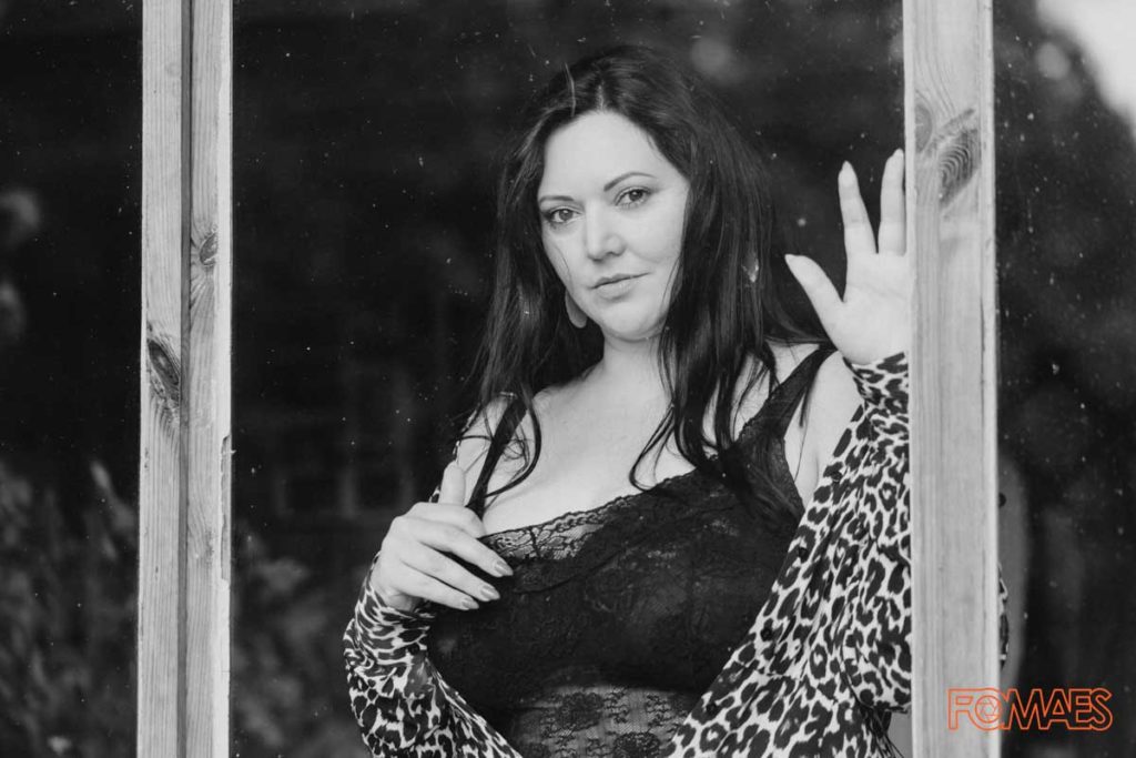 Ein S/W-Foto einer sehr hübschen Frau mit Rundungen. Die Frau ist hinter einem Fenster und stützt sich mit der linken Hand an der Fensterscheibe ab